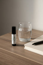 Amber glass Against the Graine roller on a nightstand or desk next to a glass of water and a journal, evoking a calming self-care moment