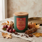 A teal green candle jar with a light wood lid and a red label reading 'Warm Apple Cider Donuts,' styled on a linen surface with autumn leaves, small pumpkins, cinnamon sticks, cloves, and red berries.