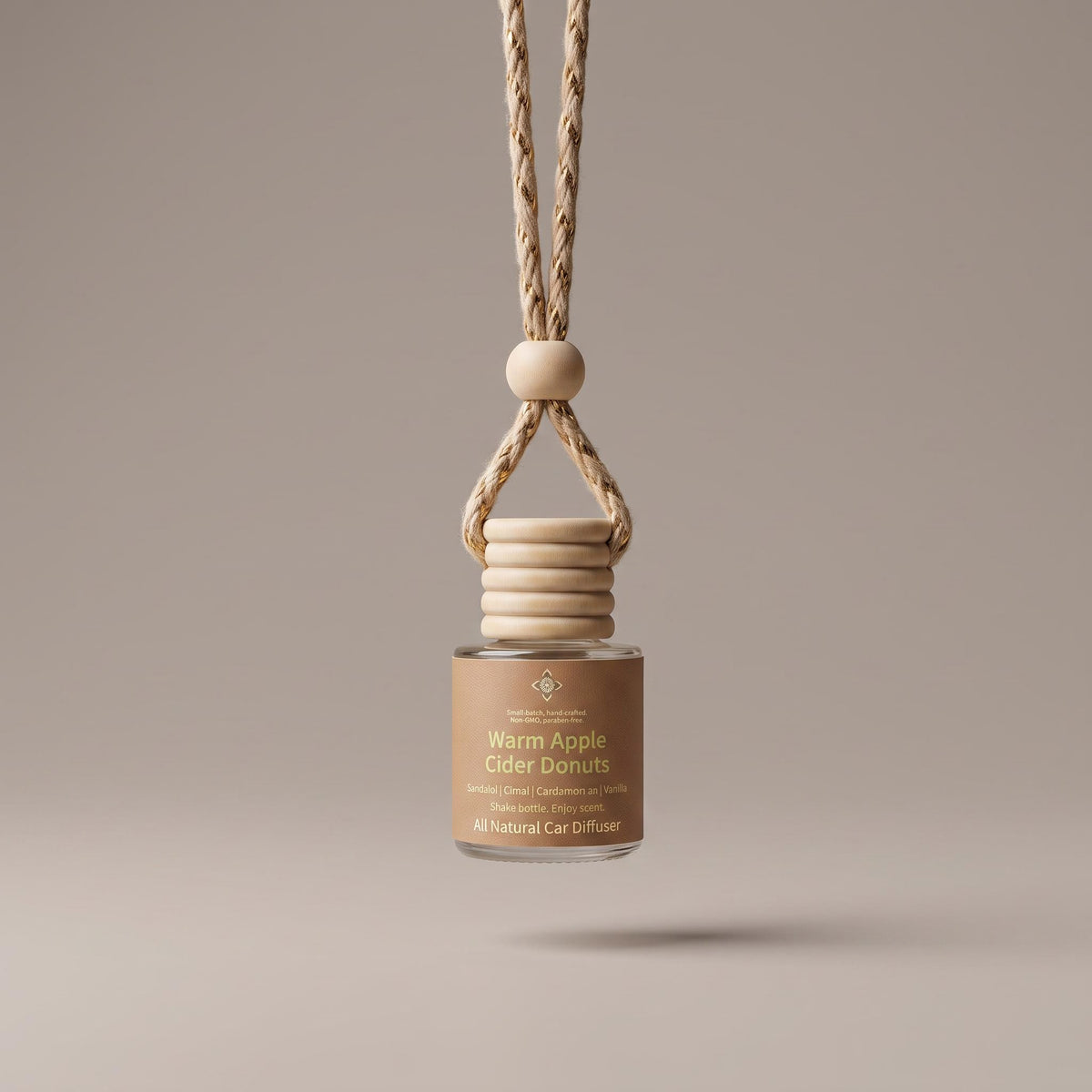 A glass car diffuser bottle with a beige label reading 'Warm Apple Cider Donuts' and a natural wood hanging collar with a braided cord, centered against a warm taupe background.