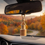 A car air diffuser bottle hanging from a rope strap under the rearview mirror inside a car, with a blurred autumn forest road scene visible through the windshield. The bottle label reads 'Warm Apple Cider Donuts' and '100% Natural Car Diffuser'.