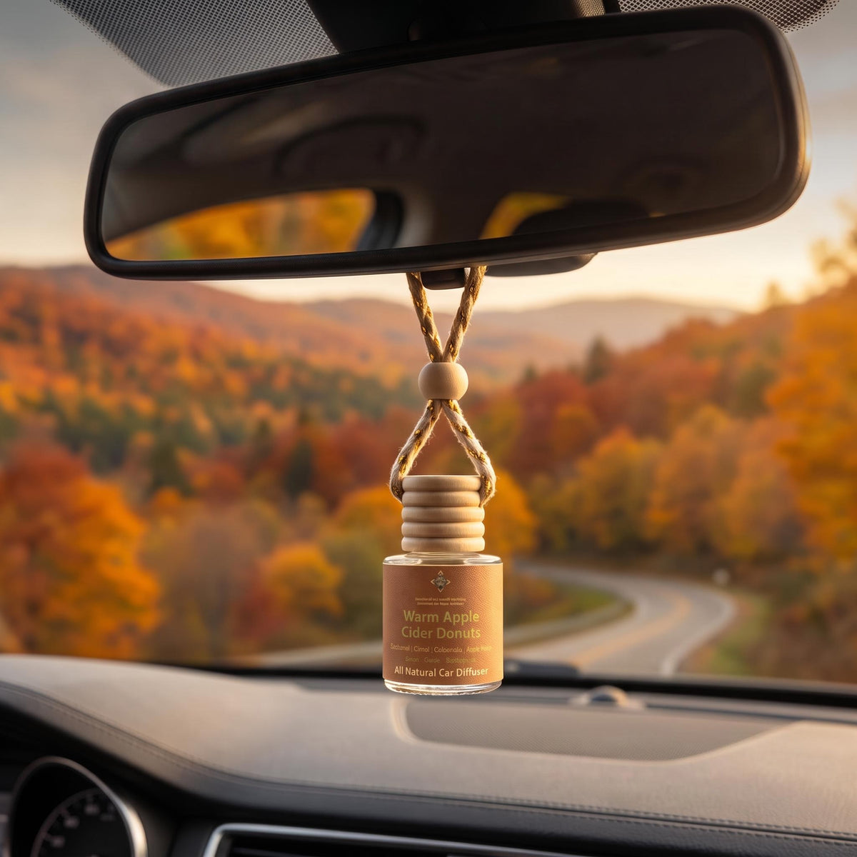 A car air diffuser bottle hanging from a rope strap under the rearview mirror inside a car, with a blurred autumn forest road scene visible through the windshield. The bottle label reads 'Warm Apple Cider Donuts' and '100% Natural Car Diffuser'.