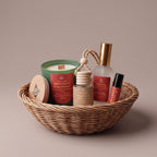 A curated gift basket set containing a green candle, a honey dipper, a frosted bottle with a gold cap, a lip balm tube, and a wooden lid, all arranged in a woven wicker basket against a solid beige background.