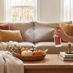 A hand holds a white spray bottle with a gold nozzle and red label, spraying a mist toward a beige sofa in a cozy living room. The scene includes autumn decor on a wooden table, including a woven tray with small pumpkins, books, and a candlestick.