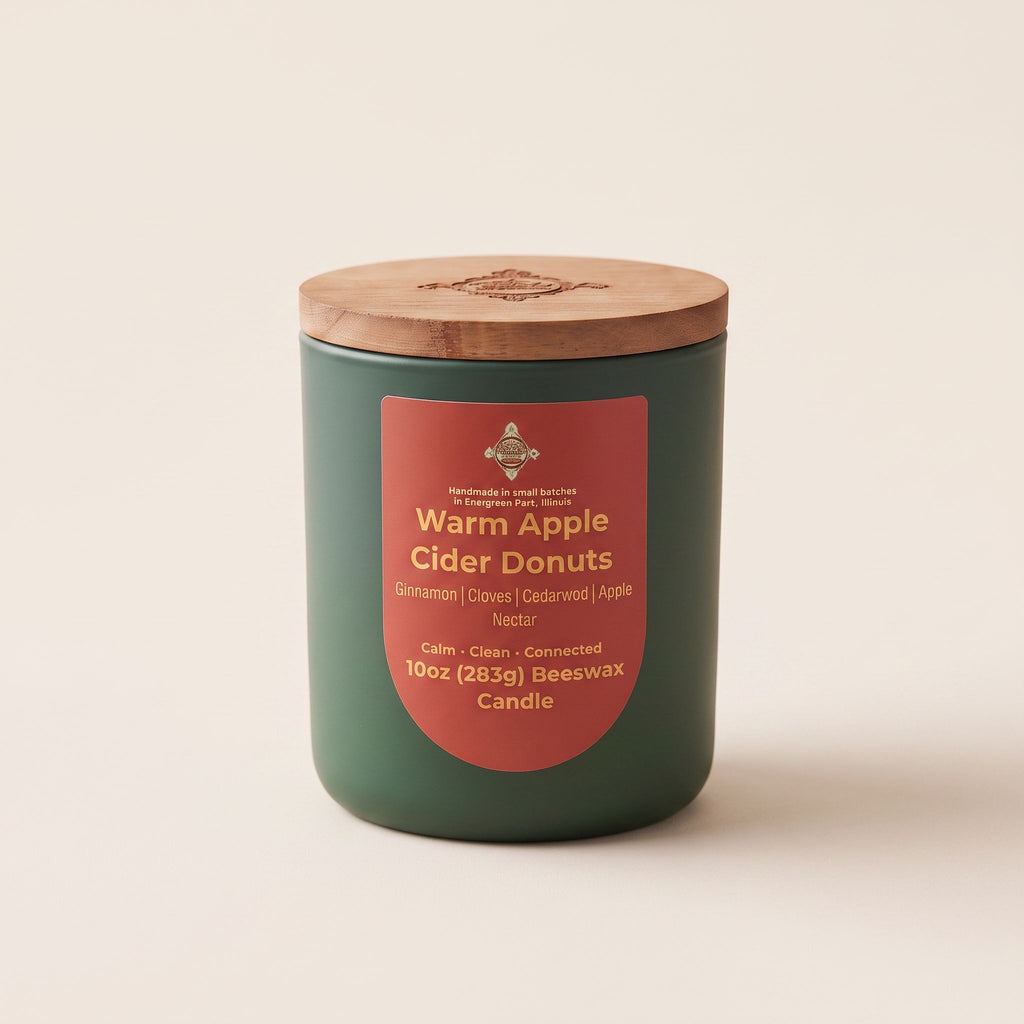 A 10oz beeswax candle in a dark green jar with a light brown wooden lid, featuring a terracotta red label that reads 'Warm Apple Cider Donuts'.