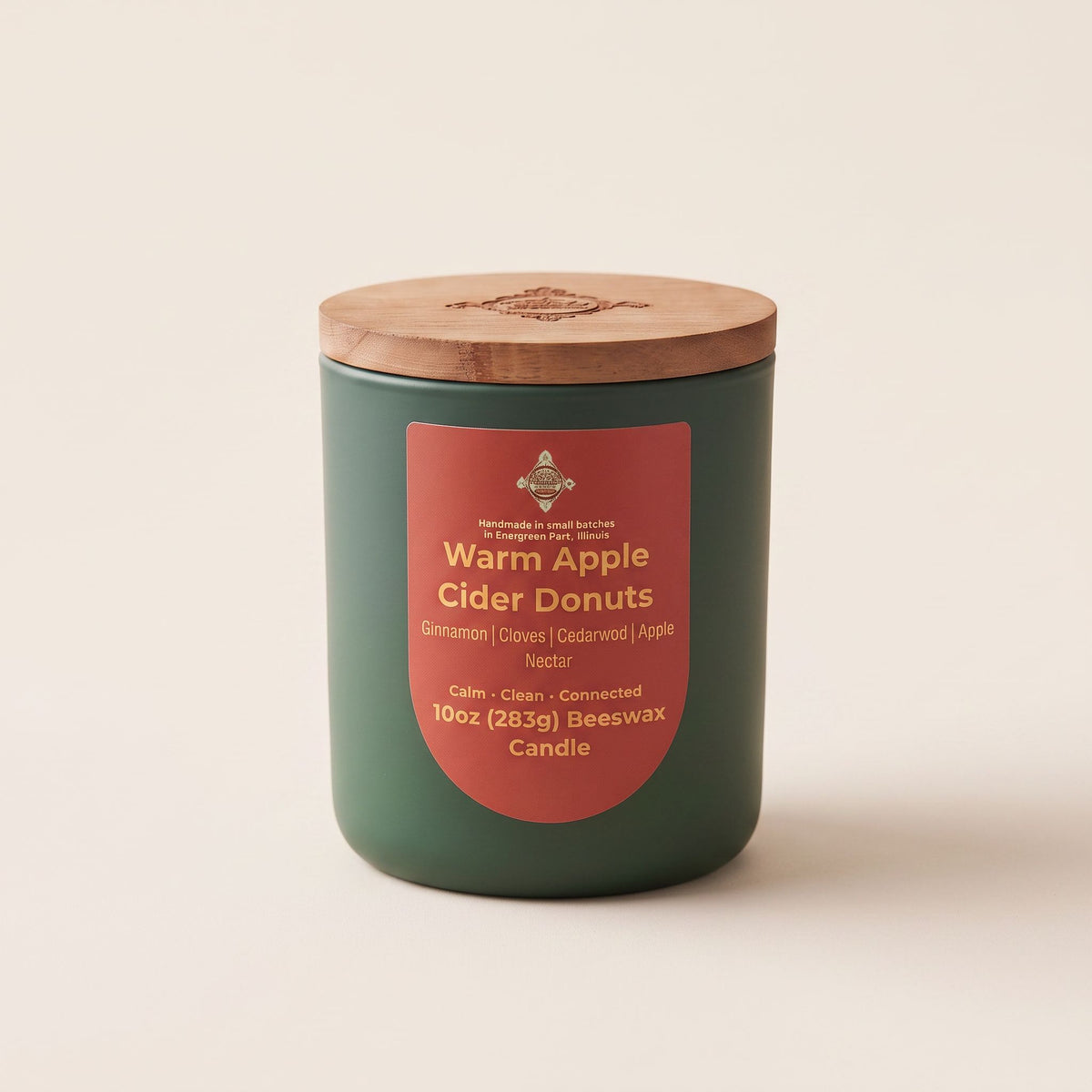 A 10oz beeswax candle in a dark green jar with a light brown wooden lid, featuring a terracotta red label that reads 'Warm Apple Cider Donuts'.