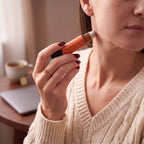 A close-up lifestyle shot of a woman applying a peach-colored body oil from a clear tube with an orange cap to her neck. She is wearing a cream cable-knit sweater and holding the product in her hand, with a softly blurred background showing a laptop and a cup of coffee.