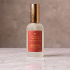 A frosted glass bottle of Warm Apple Cider Donuts room and linen spray, featuring a metallic gold cap and a terracotta-colored label, set on a polished marble surface against a textured gray background.