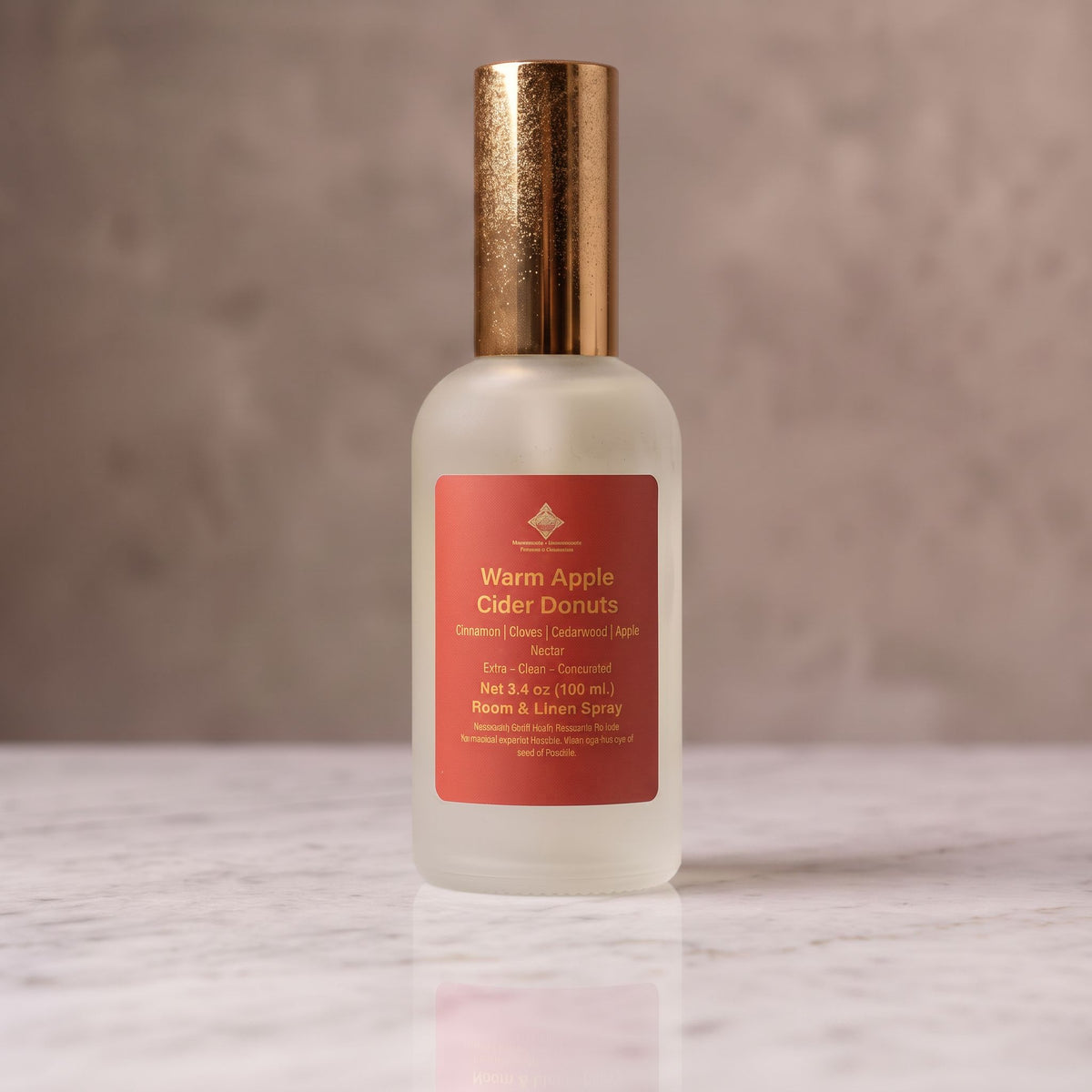A frosted glass bottle of Warm Apple Cider Donuts room and linen spray, featuring a metallic gold cap and a terracotta-colored label, set on a polished marble surface against a textured gray background.
