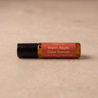 A small amber glass roller bottle with a black cap and a terracotta-colored label reading 'Warm Apple Cider Donuts' on a beige textured surface.