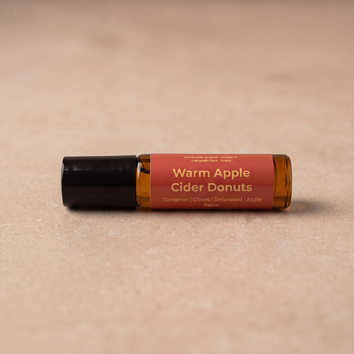 A small amber glass roller bottle with a black cap and a terracotta-colored label reading 'Warm Apple Cider Donuts' on a beige textured surface.