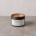 A candle in a matte black tin with a white label reading 'Distinguished Gentleman' and scent notes including Hickory, Suede, Driftwood, and Amber, photographed on a light gray textured surface against a matching neutral background.