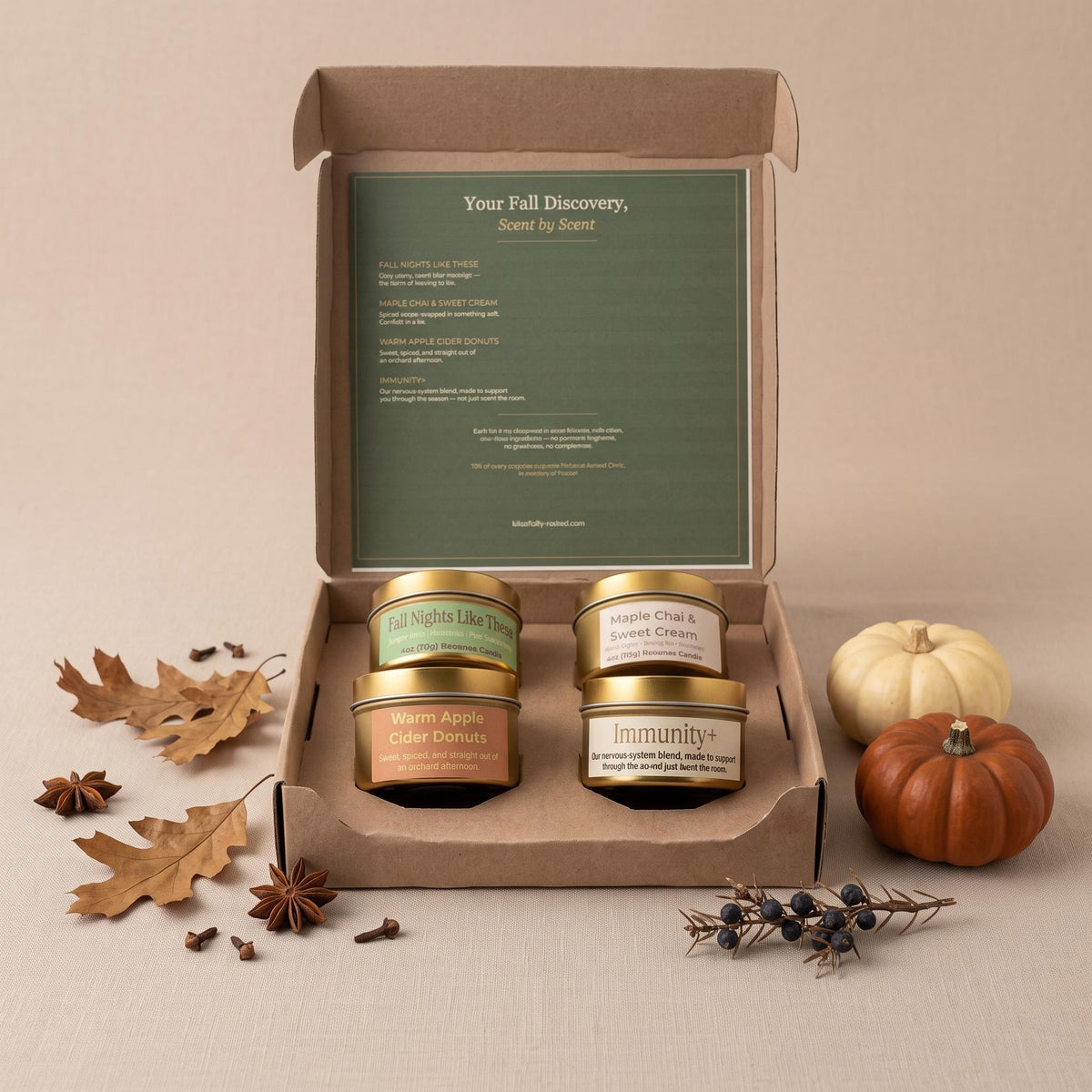 An open kraft gift box displaying four scented candles in labeled jars, with a green insert card reading 'Your Fall Discovery, Scent by Scent.' The box is styled on a beige surface with autumn props including leaves, star anise, cloves, berries, a small orange pumpkin, and a cream pumpkin.