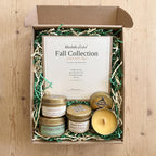 A top-down view of a 'Blissfully Perfumed Fall Collection Discovery Box' on a light wood background. The open brown cardboard box is filled with green and beige crinkle paper shred, containing a large white card and three scented candles in gold tins (Immunity, Fall Nights Like This, Maple Chai & Cream) plus one open yellow candle.