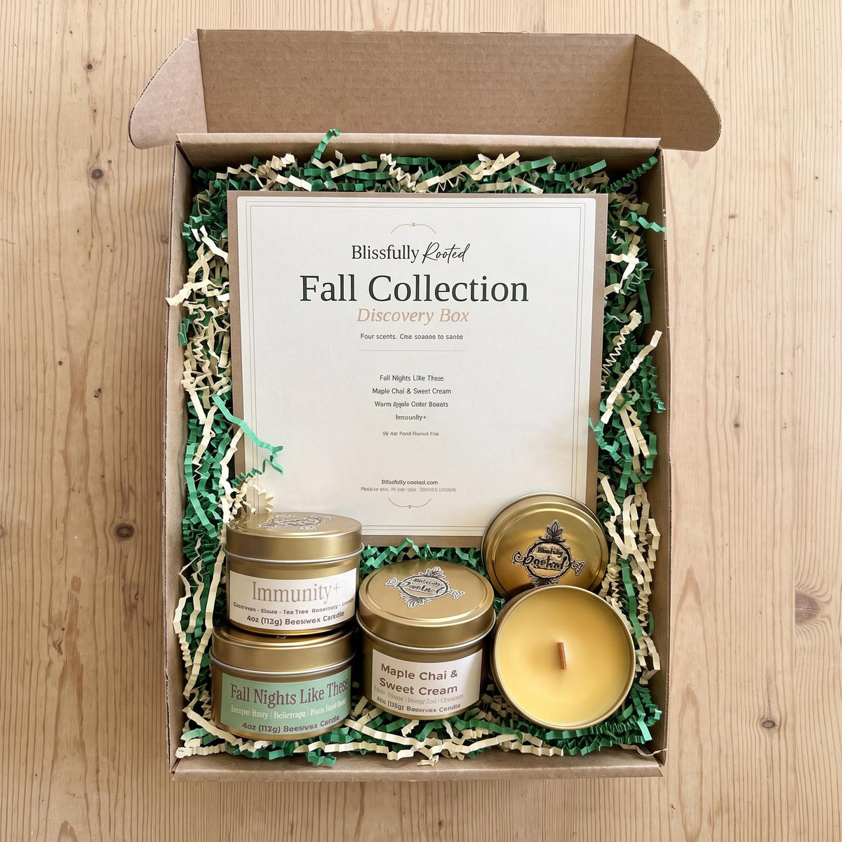 A top-down view of a 'Blissfully Perfumed Fall Collection Discovery Box' on a light wood background. The open brown cardboard box is filled with green and beige crinkle paper shred, containing a large white card and three scented candles in gold tins (Immunity, Fall Nights Like This, Maple Chai & Cream) plus one open yellow candle.