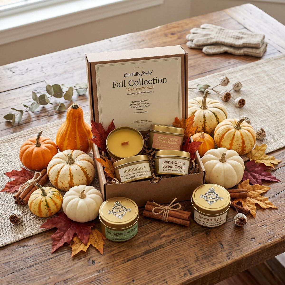 A fall-themed candle gift set displayed on a rustic wooden table. The set includes a main box labeled 'Blossom by Root Fall Collection Discovery Set' surrounded by small decorative pumpkins, fall leaves, cinnamon sticks, and several individual scented candles in glass jars with gold lids.