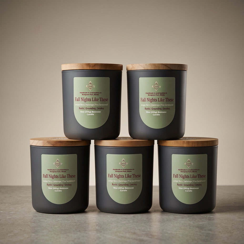 Five dark charcoal candles with light wood lids are arranged in a studio setup, with four standing on a surface and one stacked on top of the front row. Each candle has a muted sage-green label reading 'Fall Nights Like These' and smaller scent text including 'Rose & Cinnamon Clover' and 'Rose & Cedar'.