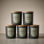 Five dark charcoal candles with light wood lids are arranged in a studio setup, with four standing on a surface and one stacked on top of the front row. Each candle has a muted sage-green label reading 'Fall Nights Like These' and smaller scent text including 'Rose & Cinnamon Clover' and 'Rose & Cedar'.