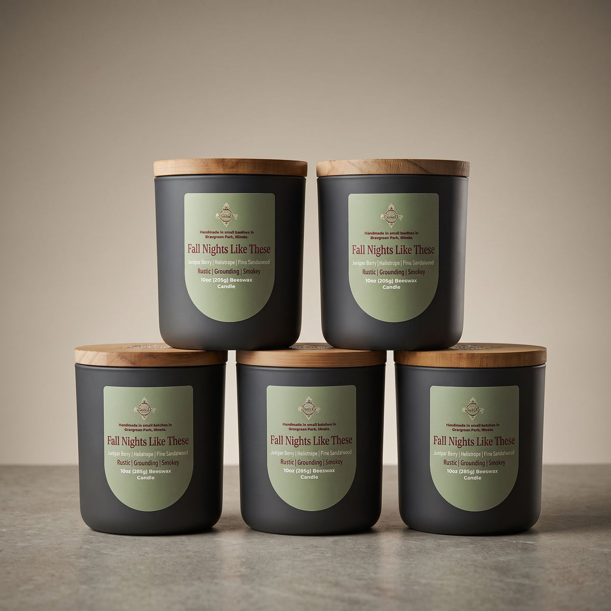 Five dark charcoal candles with light wood lids are arranged in a studio setup, with four standing on a surface and one stacked on top of the front row. Each candle has a muted sage-green label reading 'Fall Nights Like These' and smaller scent text including 'Rose & Cinnamon Clover' and 'Rose & Cedar'.
