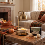 A brass table lamp with a curved neck and dome shade stands on a round base on a rustic wooden coffee table in a cozy living room. A bowl of yellow flowers, a small orange pumpkin, and a box of fall night lights sit on the table alongside a plaid throw blanket and fireplace in the background.