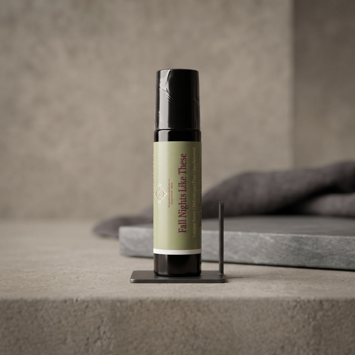 A small cylindrical bottle labeled 'Fall Nights Like These' stands upright on a black display stand against a textured gray background with a folded dark gray cloth behind it.