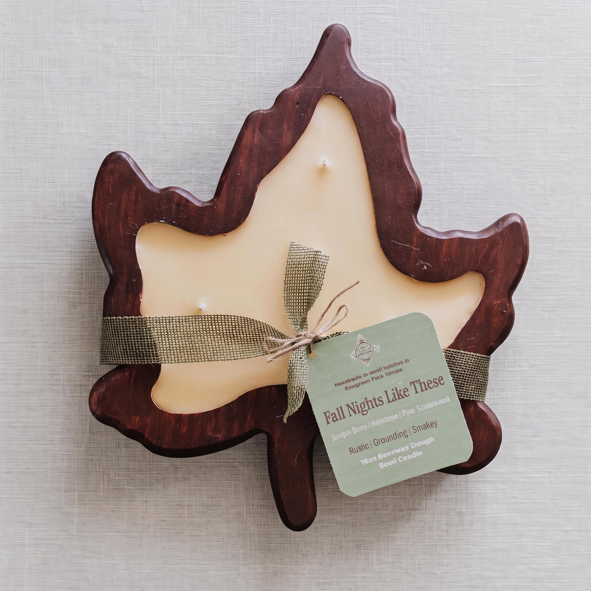 A leaf-shaped candle with a dark reddish-brown wooden frame and a cream-colored wax center, tied with a rustic olive ribbon and a green paper tag that reads 'Fall Nights Like These.' The candle is photographed on a light neutral fabric background.