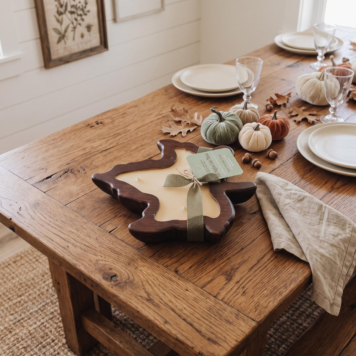 A rustic wooden table set for dining, featuring a central autumn-themed candle in a leaf-shaped wooden holder with a sage green ribbon and tag. The table is styled with white plates, wine glasses, small pumpkins, and scattered fall leaves on a warm wood surface.