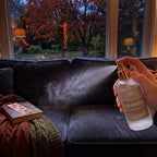 A hand holds a frosted glass room spray bottle with a gold spray nozzle, spraying mist onto a dark couch in a cozy living room setting with a window showing autumn trees outside.