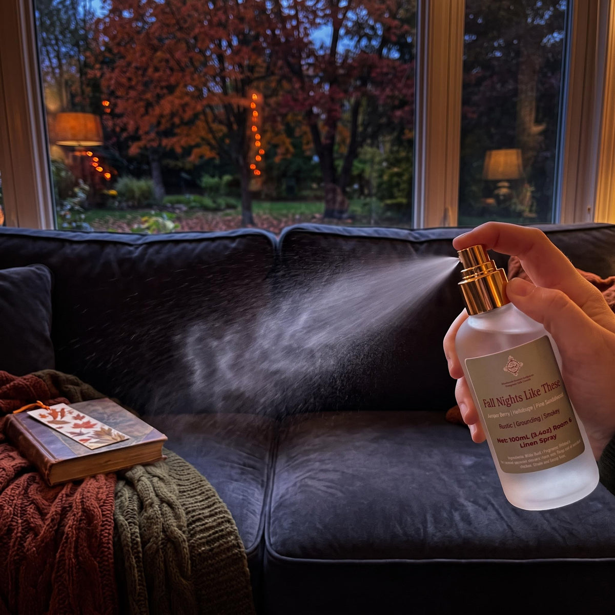 A hand holds a frosted glass room spray bottle with a gold spray nozzle, spraying mist onto a dark couch in a cozy living room setting with a window showing autumn trees outside.