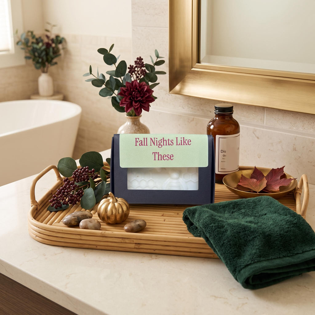 A boxed bath or self-care product sits on a bamboo tray in a bathroom setting, surrounded by a folded dark green towel, decorative leaves, a small pumpkin, stones, and an amber bottle. The front of the box has a light green label reading 'Fall Nights Like These.