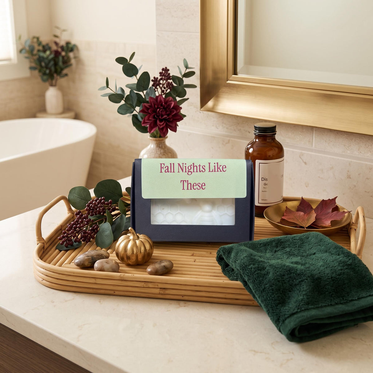 A boxed bath or self-care product sits on a bamboo tray in a bathroom setting, surrounded by a folded dark green towel, decorative leaves, a small pumpkin, stones, and an amber bottle. The front of the box has a light green label reading 'Fall Nights Like These.