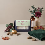 A boxed candle set styled on a beige tabletop with eucalyptus, a small gold pumpkin, stones, a dark green towel, and a brown amber bottle. The main product is a dark navy rectangular candle box with a light green label reading 'Fall Nights Like These' and a clear window showing a white honeycomb-patterned candle inside.