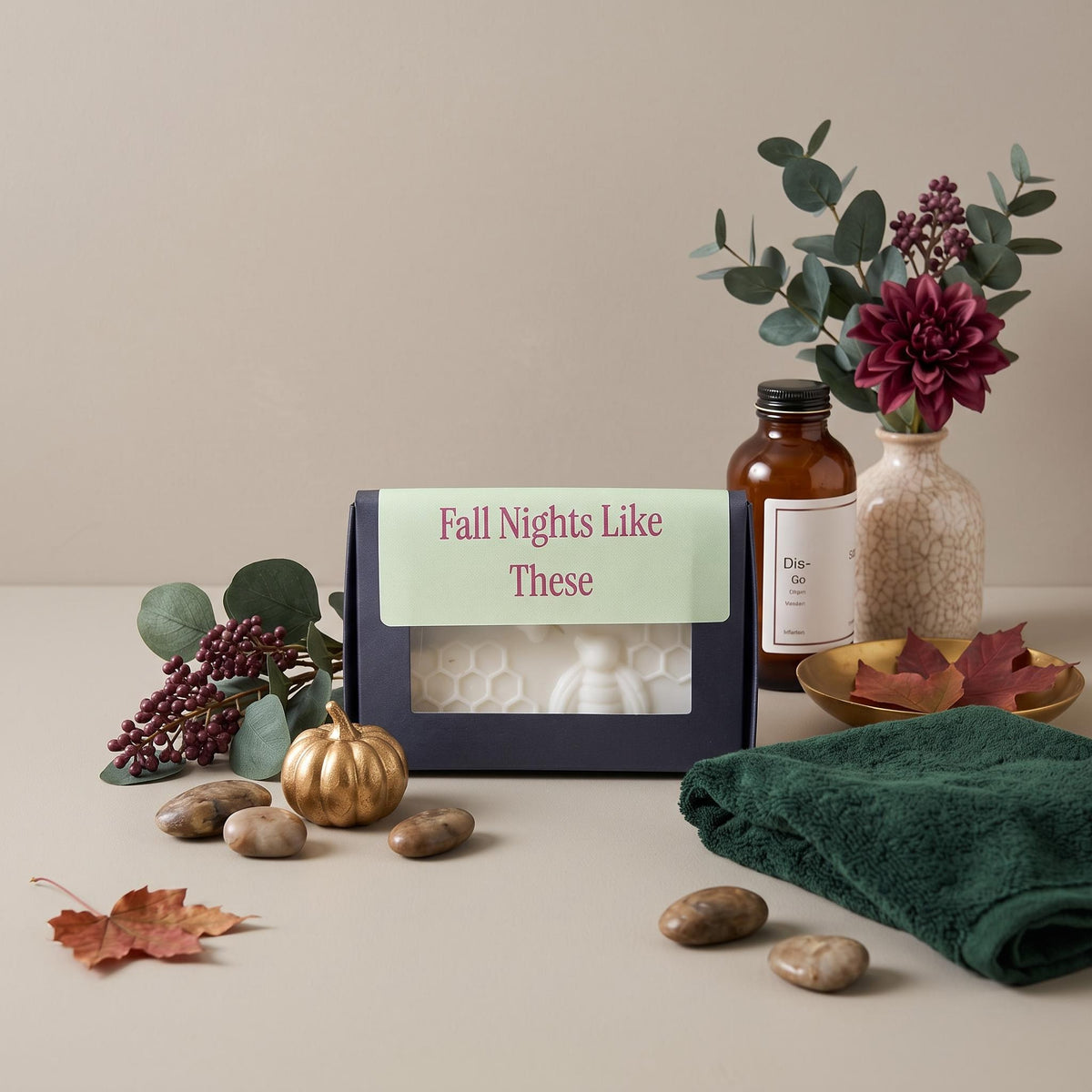 A boxed candle set styled on a beige tabletop with eucalyptus, a small gold pumpkin, stones, a dark green towel, and a brown amber bottle. The main product is a dark navy rectangular candle box with a light green label reading 'Fall Nights Like These' and a clear window showing a white honeycomb-patterned candle inside.