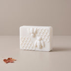 A rectangular white soap bar featuring embossed bees and honeycomb patterns, set on a neutral beige surface with a small dried leaf in the corner.