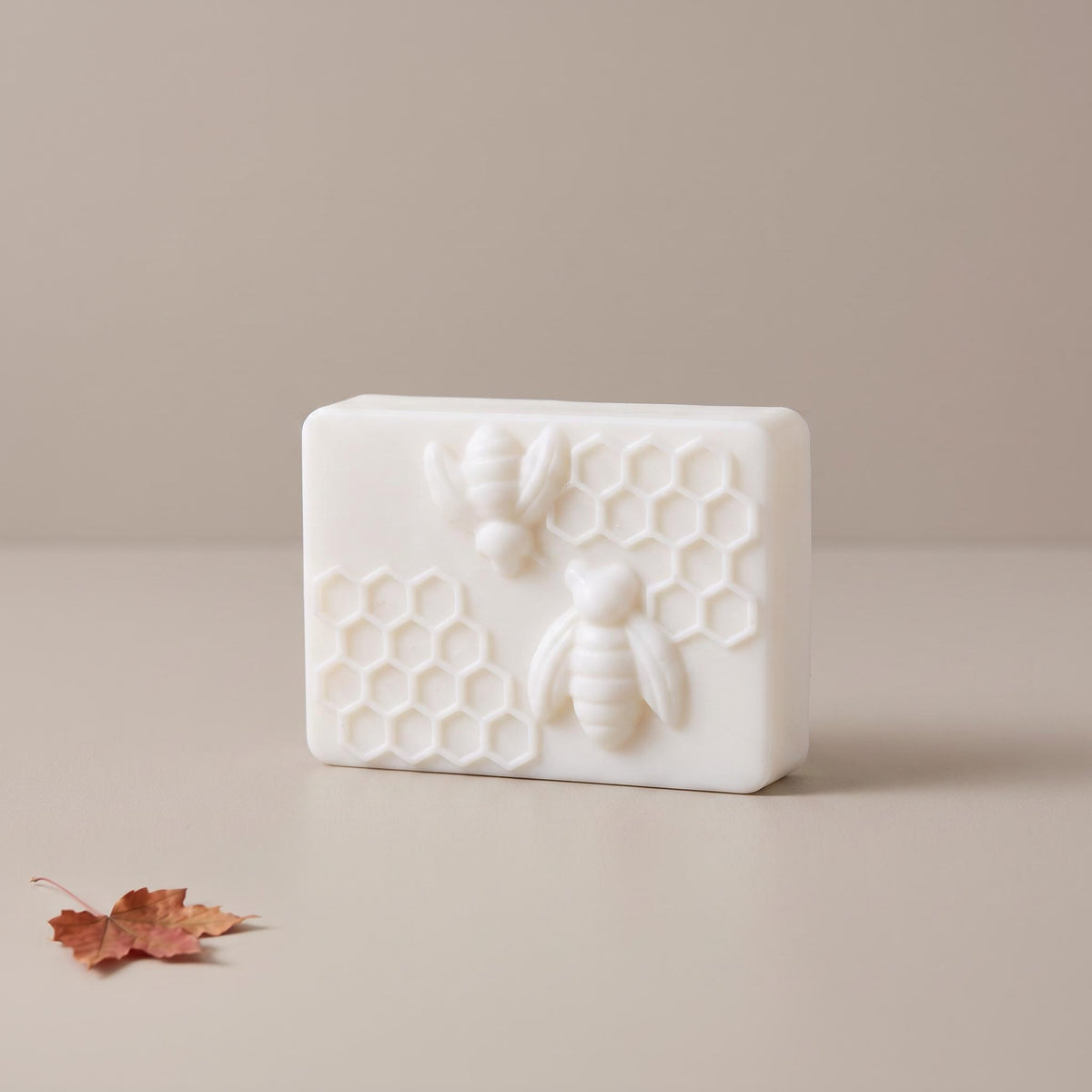 A rectangular white soap bar featuring embossed bees and honeycomb patterns, set on a neutral beige surface with a small dried leaf in the corner.