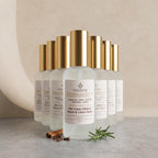 Six frosted glass room and linen spray bottles with shiny gold caps arranged in a row on a light stone surface. The front bottle is labeled 'Immunity+' and shows rosemary and cinnamon props at its base against a soft beige background.