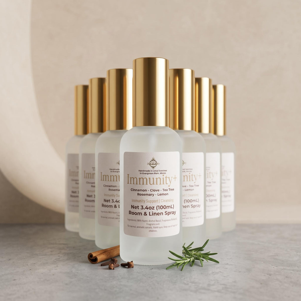 Six frosted glass room and linen spray bottles with shiny gold caps arranged in a row on a light stone surface. The front bottle is labeled 'Immunity+' and shows rosemary and cinnamon props at its base against a soft beige background.