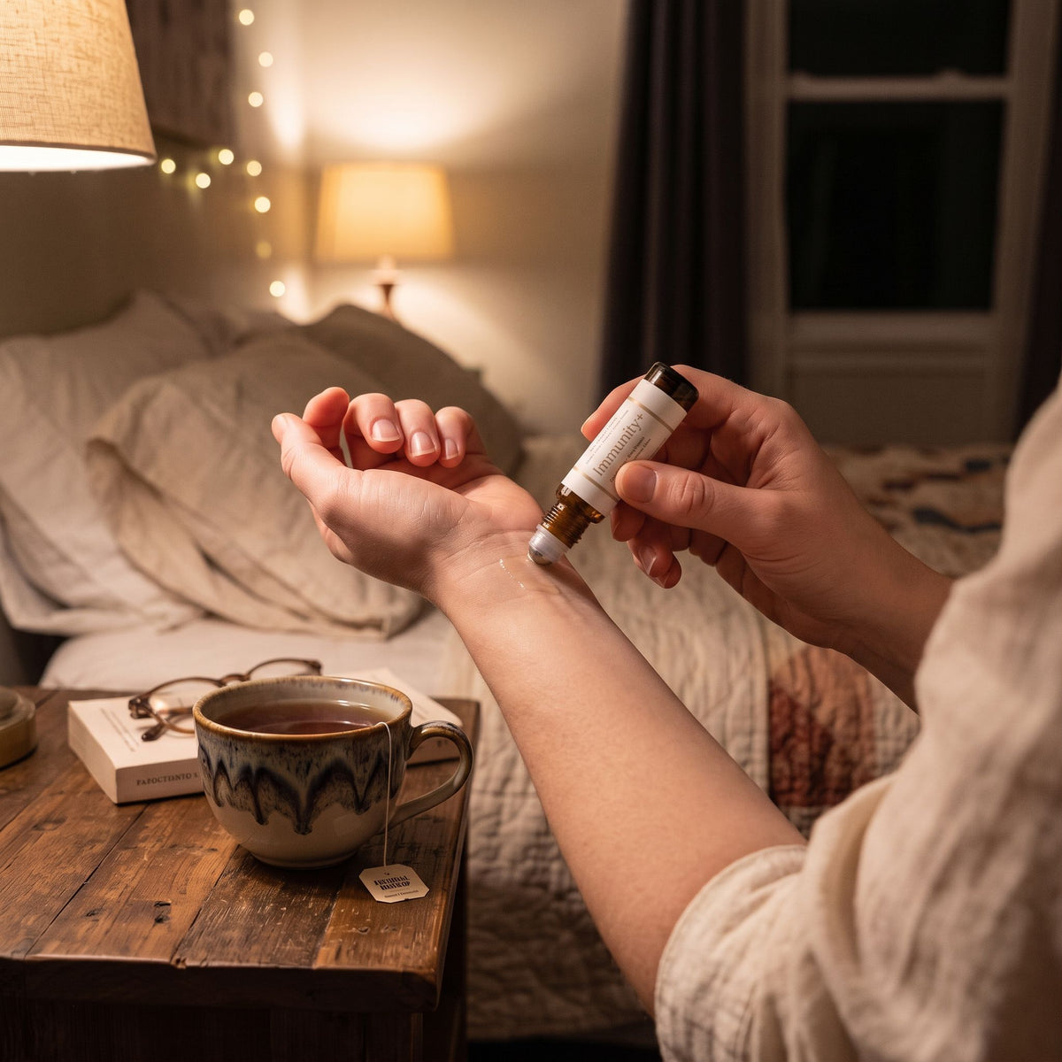 A person applies a small essential oil or serum dropper bottle to their inner wrist in a cozy bedroom setting. A ceramic mug, glasses, and a book sit on a wooden bedside table in the foreground, with a bed and warm string lights in the background.