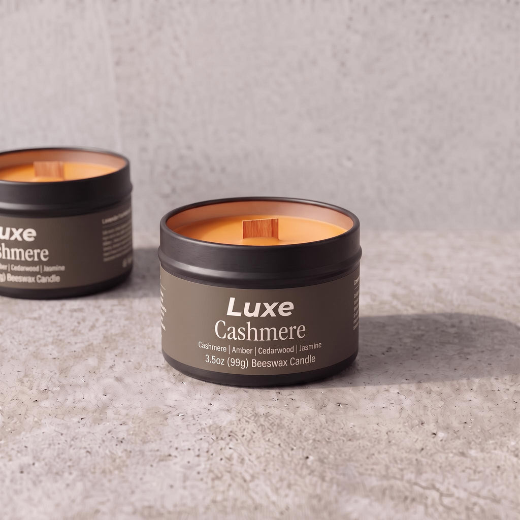 Two LuxE Cashmere beeswax candles in dark metal tins on a light gray textured surface, with one candle in the foreground in sharp focus and the other blurred in the background.