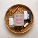 A curated collection of spa and self-care items arranged on a round woven wicker tray, including a Maple Chai & Sweet Cream scented candle, a matching hand and body spray, a small black box of wax melts, and a lavender oil roller, styled with dried orange slices and cinnamon sticks on a textured off-white background.