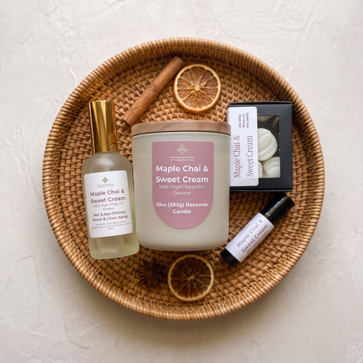 A curated collection of spa and self-care items arranged on a round woven wicker tray, including a Maple Chai & Sweet Cream scented candle, a matching hand and body spray, a small black box of wax melts, and a lavender oil roller, styled with dried orange slices and cinnamon sticks on a textured off-white background.