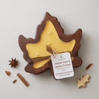 A maple leaf-shaped bowl candle with a dark brown wooden frame and pale yellow wax, tied with a brown ribbon and a white label reading 'Maple Chai & Sweet Cream.' The candle is photographed on a light neutral background with scattered cinnamon sticks, star anise, and dried botanicals around it.