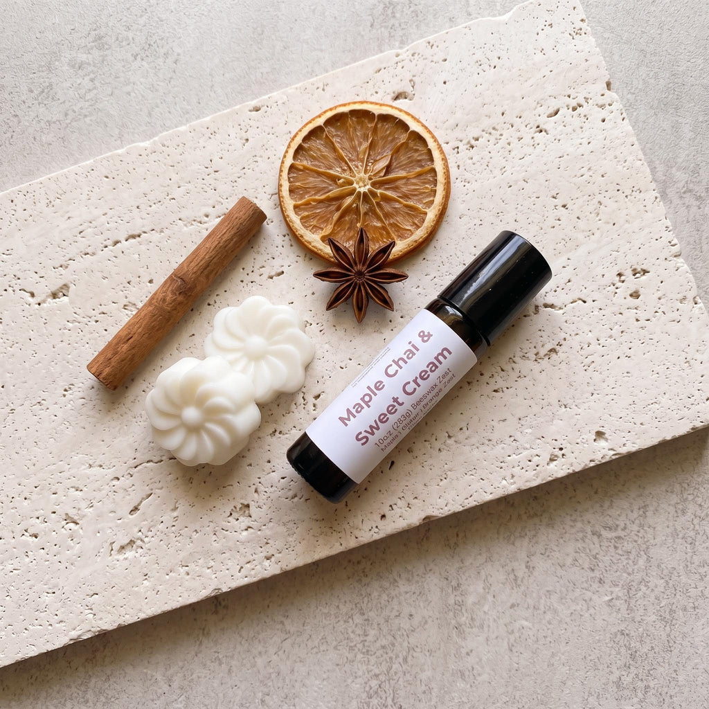 A small black roll-on bottle labeled 'Maple Chai & Sweet Cream' is arranged on a light stone slab with a cinnamon stick, dried orange slice, star anise, and two white wax melts.