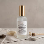A frosted glass room and linen spray bottle with a metallic gold cap, labeled 'Maple Chai & Sweet Cream', set on a textured neutral background with dried botanicals and a spoon.