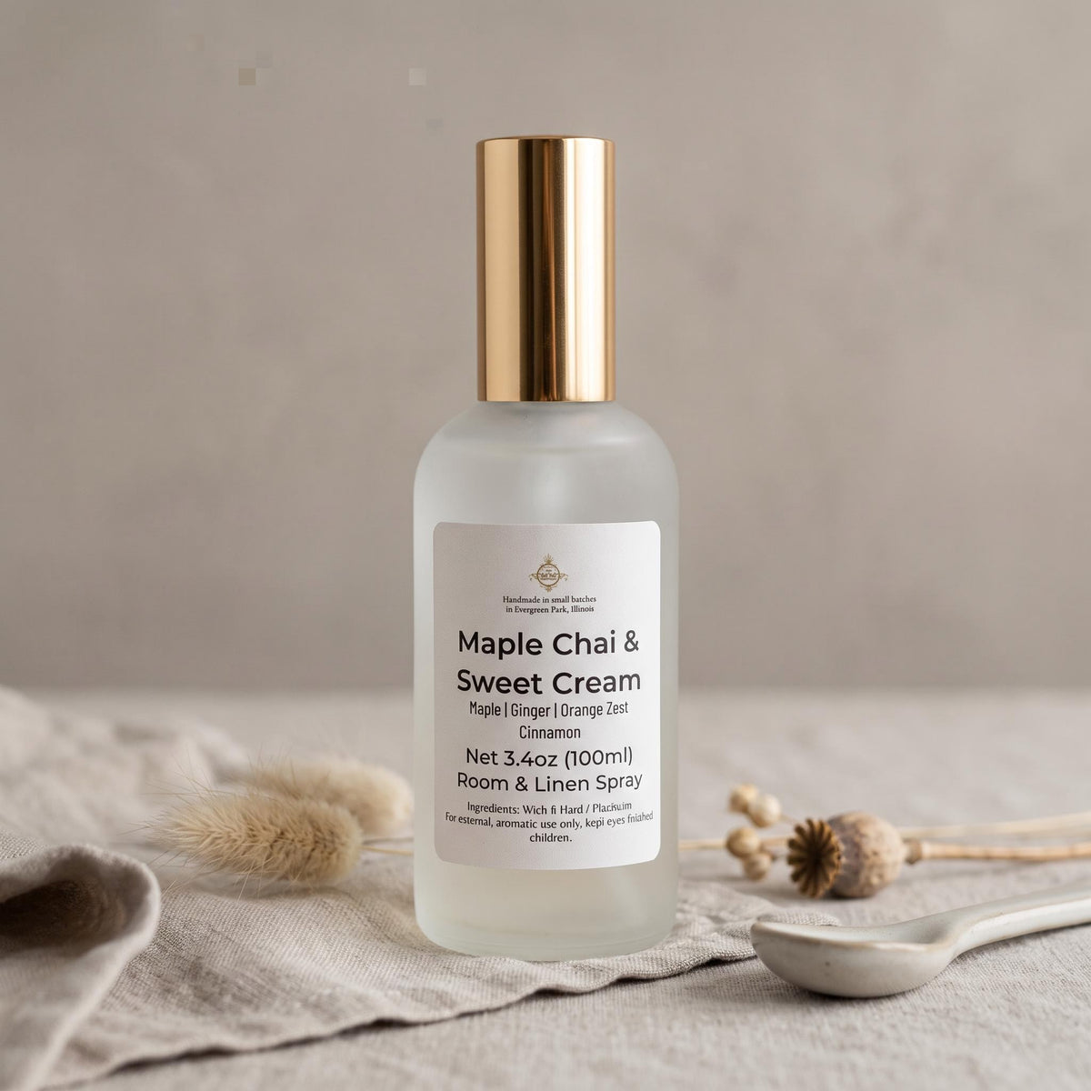 A frosted glass room and linen spray bottle with a metallic gold cap, labeled 'Maple Chai & Sweet Cream', set on a textured neutral background with dried botanicals and a spoon.