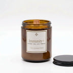 An amber glass jar candle with a white label reading 'Immunity+' and a wooden wick, shown on a white background with its black lid placed to the right.