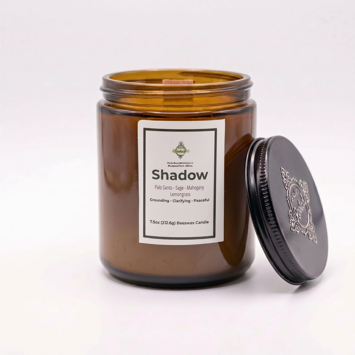 An amber glass jar candle with a white label reading 'Shadow' and a black metal lid leaning against the jar on a clean white background.