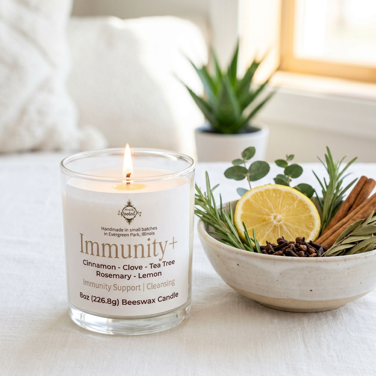 A lit white candle in a clear glass jar labeled 'Immunity+' sits on a light surface beside a bowl of fresh rosemary, cinnamon sticks, cloves, and a lemon slice. The background is softly blurred with a plant and window light.
